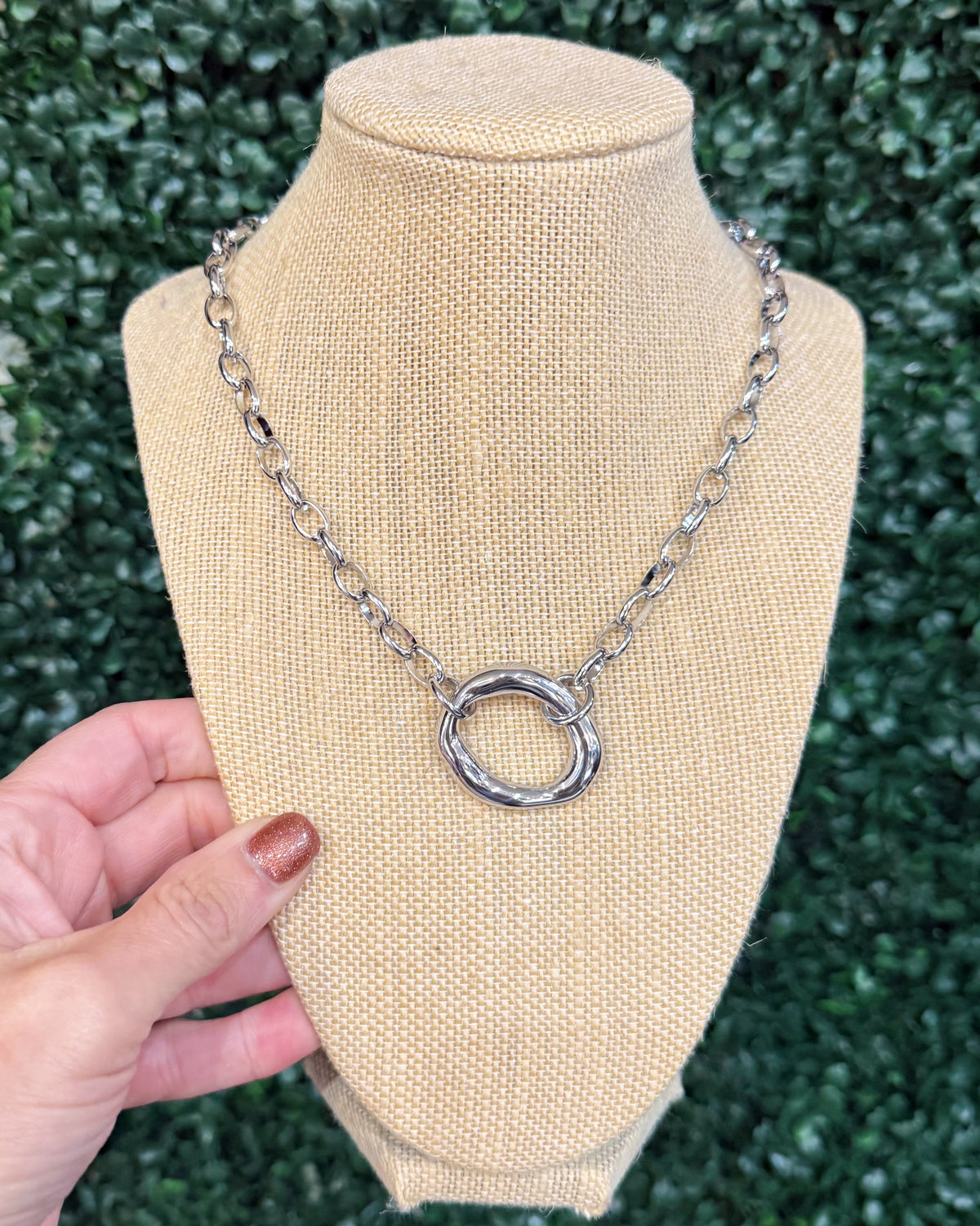 A silver-toned chain necklace with a textured circular ring pendant, displayed on a beige fabric jewelry bust. A hand with brown nail polish is holding the left side of the bust, and a green hedge wall serves as the background.