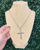 A silver cross pendant necklace displayed on a beige burlap-covered jewelry bust, with a hand holding the left side of the bust against a green leafy background.