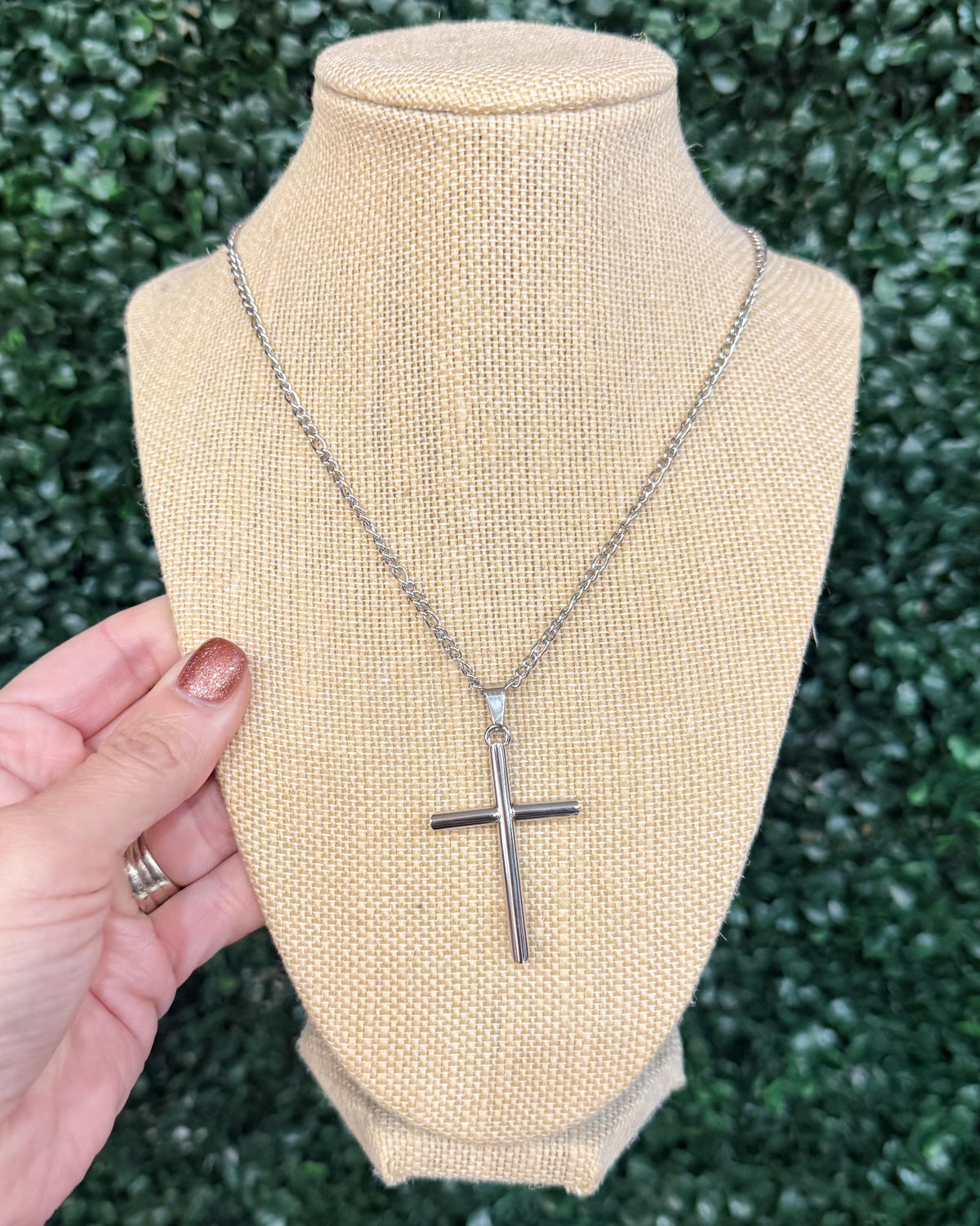 A silver cross pendant necklace displayed on a beige burlap-covered jewelry bust, with a hand holding the left side of the bust against a green leafy background.