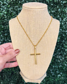 A gold cross pendant necklace displayed on a beige burlap jewelry bust, with a hand holding the left side of the bust. The background is a blurred green hedge.