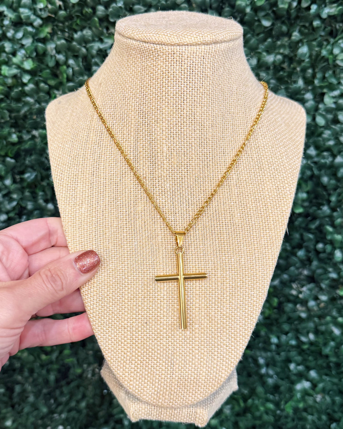 A gold cross pendant necklace displayed on a beige burlap jewelry bust, with a hand holding the left side of the bust. The background is a blurred green hedge.