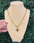 A gold chain necklace with a cross pendant displayed on a beige fabric jewelry bust, with a hand holding the left side of the bust against a green leaf background.