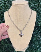 A silver-toned cross pendant necklace displayed on a beige fabric mannequin bust, with a hand holding the left side of the bust for scale, against a background of green foliage.