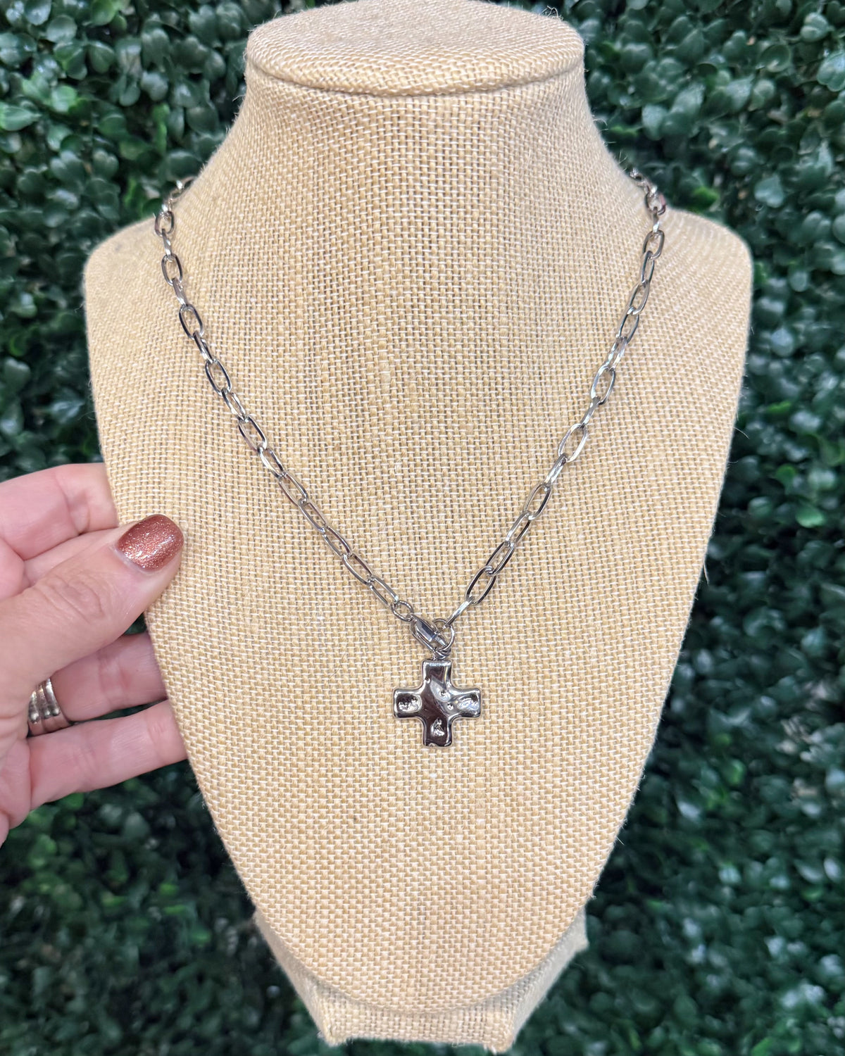 A silver-toned cross pendant necklace displayed on a beige fabric mannequin bust, with a hand holding the left side of the bust for scale, against a background of green foliage.