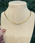A gold chain necklace with three small clear stones, displayed on a beige fabric jewelry bust. A hand with glittery pink nail polish is visible on the left side, holding the bust in front of a green leafy background.