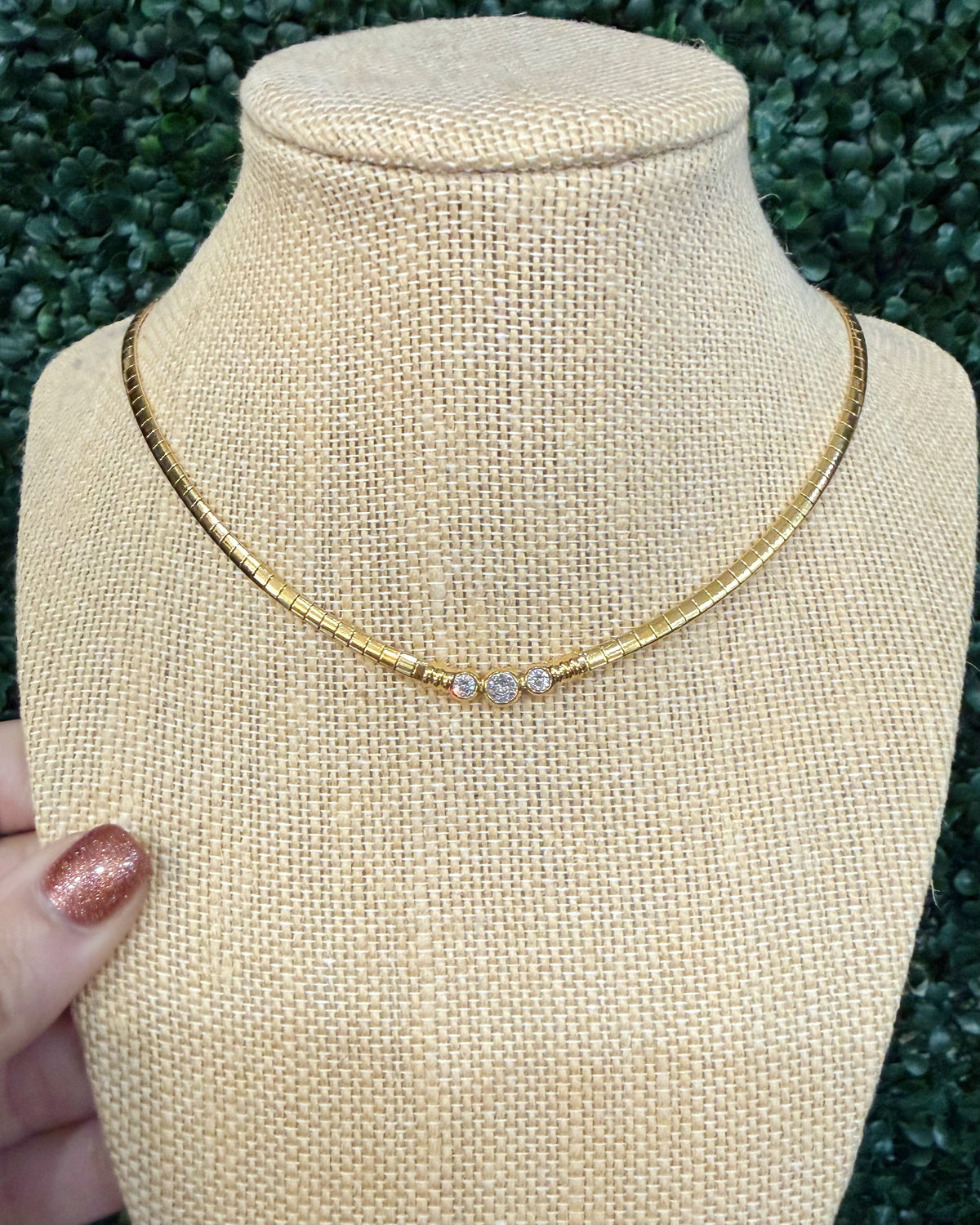 A gold chain necklace with three small clear stones, displayed on a beige fabric jewelry bust. A hand with glittery pink nail polish is visible on the left side, holding the bust in front of a green leafy background.