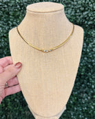 A gold chain necklace with three small clear stones, displayed on a beige fabric jewelry bust. A hand with glittery nail polish is holding the side of the bust, and a green hedge is visible in the background.