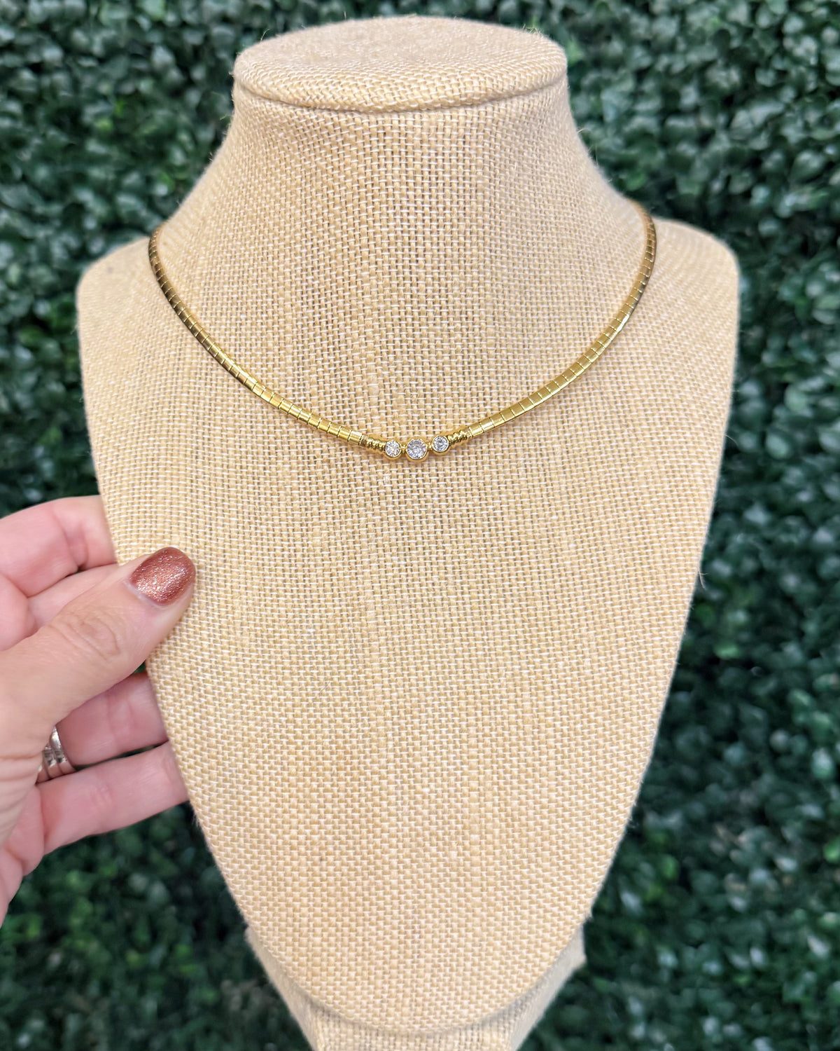 A gold chain necklace with three small clear stones, displayed on a beige fabric jewelry bust. A hand with glittery nail polish is holding the side of the bust, and a green hedge is visible in the background.