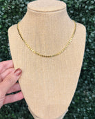 A gold chain necklace displayed on a beige fabric jewelry bust, with a hand holding the left side of the bust. The background is a dark green leafy hedge.