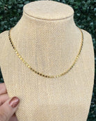A delicate gold chain necklace displayed on a beige woven jewelry bust, with a hand holding the left side of the bust. The background consists of blurred green foliage.