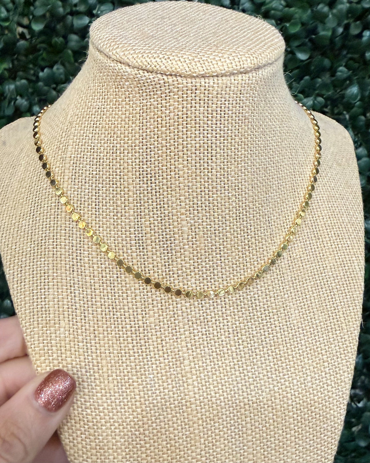 A delicate gold chain necklace displayed on a beige woven jewelry bust, with a hand holding the left side of the bust. The background consists of blurred green foliage.