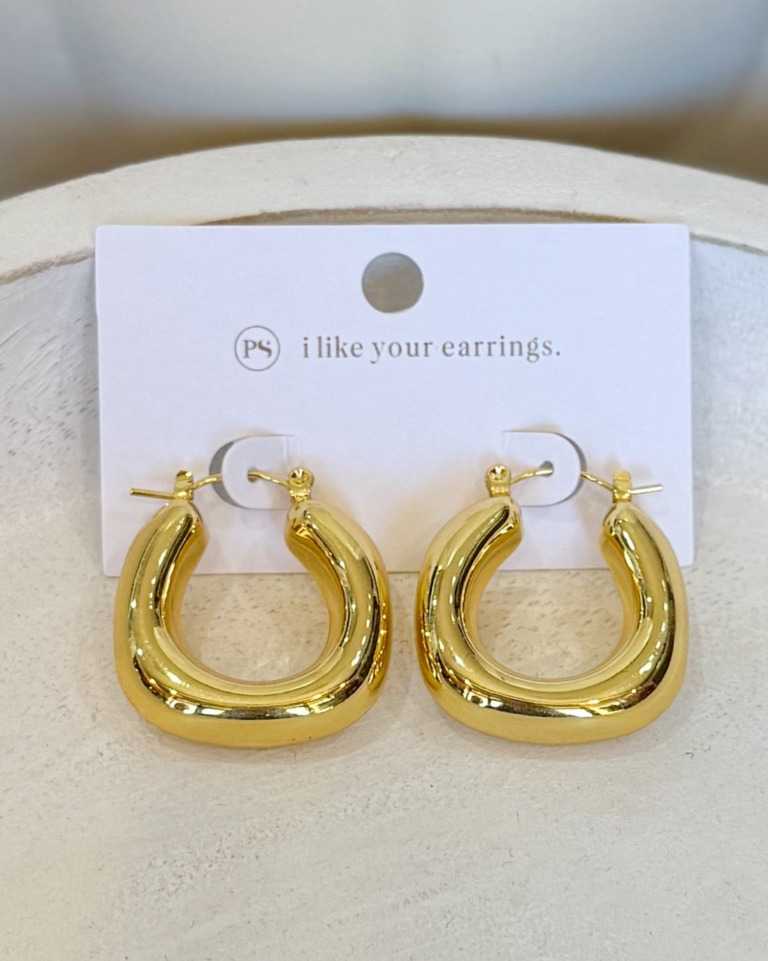 A pair of chunky, polished gold hoop earrings displayed on a white card that reads 'PS i like your earrings.' against a neutral, light beige textured background.